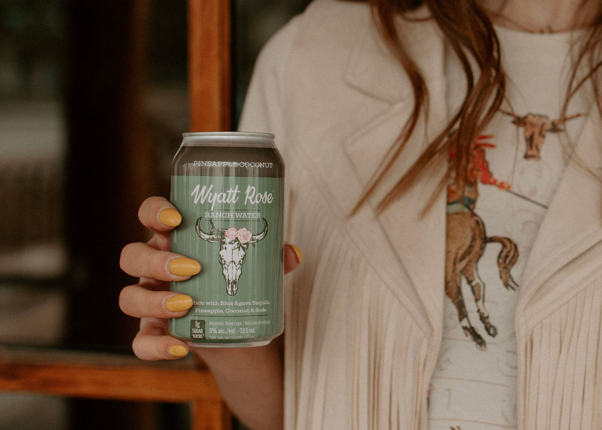 Close up of a woman in a fringe jacket and cowboy motif holding a can of Wyatt Rose Ranch Water